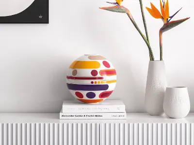 A colourful La Boule Miami standing on books next to white vases and bird of paradise flowers on a white shelf.