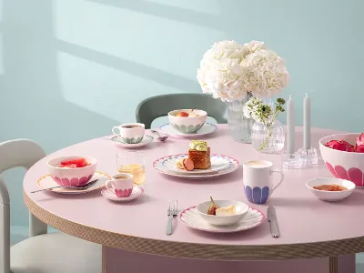 Ein runder Tisch mit Tellern, Tassen und Schüsseln voller Essen, dekoriert mit rosa Mustern. In der Mitte stehen eine Vase mit weißen Blumen und zwei unbeleuchtete Kerzen. Stühle umgeben den Tisch.