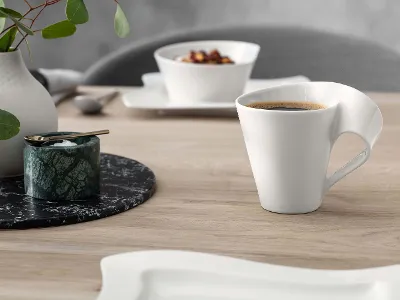 A modern white coffee cup with a striking handle design, next to a small container and a vase with green leaves on a wooden table.