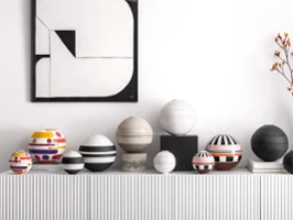 Eine Reihe gemusterter kugelförmiger Geschirrsets auf einem weißen Regal, darunter ein geometrisches Schwarz-Weiß-Gemälde mit einer Vase mit orangefarbenen Blumen.