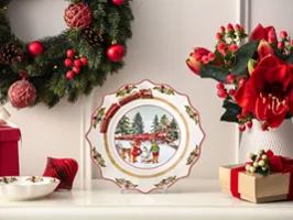 Ein dekorativer Weihnachtsteller mit einer Winterszene wird auf einem Kaminsims mit Weihnachtskränzen, Blumen und eingepackten Geschenken präsentiert.