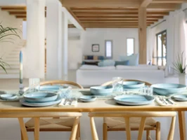 A dining table set with blue Perlemor Aqua tableware and glasses in a bright, modern, open-plan living area with wooden beams.