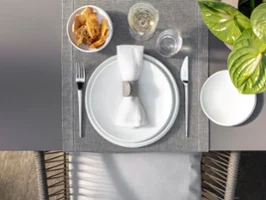 Vue de dessus d'un couvert avec des assiettes blanches NewMoon, une serviette, des couverts, un bol à céréales, un verre et une plante sur un set de table tissé.