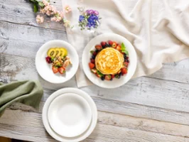 Ein Stapel Pfannkuchen mit Beeren wird auf einem Teller von Villeroy & Boch New Cottage serviert, auf einem anderen Teller gibt es geschnittenes Obst. Der Holztisch ist mit Blumen und einer drapierten Tischdecke geschmückt.