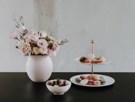 Eine weiße Vase mit rosa Blumen, ein mehrstöckiges Tablett mit Desserts und eine Schale mit Pralinen auf einem dunklen Tisch.
