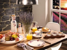 Un couvert pour le petit-déjeuner avec la vaisselle de Villeroy & Boch Artesano Provençal Lavande Un couvert de petit-déjeuner avec la vaisselle Villeroy & Boch Artesano Provençal Lavande, orné de fruits, d'un croissant, de céréales et de jus d'orange. Un vase de lavande trône élégamment au centre de la table. La fenêtre offre une vue pittoresque d'un coucher de soleil sur un champ de fleurs violettes.