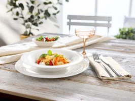 Une assiette de pâtes avec des légumes et des herbes, une salade et une boisson sur une table en bois dressée pour une personne.