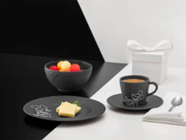 Black tableware from the Manufacture Mickey collection next to a white gift box Black tableware from the Manufacture Mickey collection with fruit, buttered bread and coffee, alongside a white gift box, napkin and spoon on a divided background.