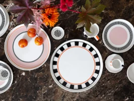 Servizio da tavola Memphis di Villeroy & Boch con piatti decorativi, una tazza, tazzine e prugne su una superficie in marmo marrone decorata con fiori luminosi.
