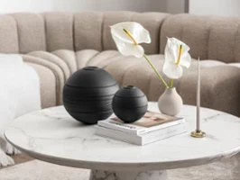 The Villeroy & Boch black La Boule, a beige vase with white flowers, a candleholder and some books in a beige living room setting.