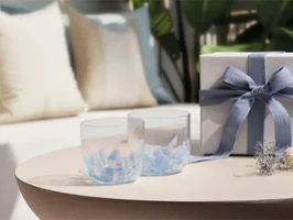 Two clear Like Aqua glasses and a white gift box with a blue ribbon on a round table; sofa and cushions in the background.