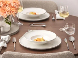 Tavolo da pranzo apparecchiato della collezione Afina Una tavola ben apparecchiata della collezione Afina con due piatti con piatti gourmet, bicchieri da vino, bicchieri da acqua, posate d'argento, tovaglioli di stoffa e un vaso con fiori di pesco su una superficie di legno.