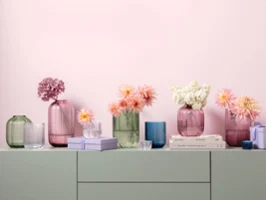 Various glass vases in pastel colors from the Fleur collection, decorated with flowers, are arranged on a light surface with books and small decorative items in front of a pink wall.