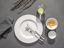Piemont cutlery on a plate A white plate with fork and knife, a glass of water with lemon, a bowl of crackers and a napkin on a grey table.