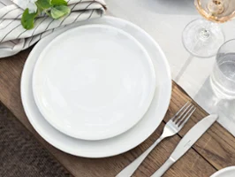 Un Artesano- set de table de Villeroy & Boch avec des assiettes blanches Un Artesano- set de table de Villeroy & Boch avec des assiettes blanches, des couverts, une serviette rayée avec un motif de fleurs, un verre de rosé, du pain sur une planche et un bol avec des artichauts.