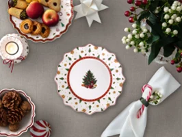 Ein festlich gedeckter Tisch mit einem dekorierten Teller mit Weihnachtsbaummotiv, Keksen, Obst, Kerzen, Servietten und einem Blumengesteck.