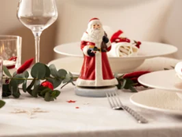 Eine festlich gedeckte Tafel mit einer Nostaligic Melody Weihnachtsmann Spieluhr in der Mitte, umgeben von Tellern, Gläsern, Besteck und Grünpflanzen.