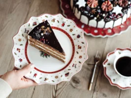 One hand holds a plate with a piece of chocolate cake. The table also features a decorated whole cake, a cup of coffee, a fork and a saucer, and plate set from Toy’s Delight.