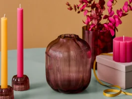 Vasi in vetro scanalato Fleur e candele colorate Primo piano di vasi in vetro scanalato nei toni del mirtillo, con candele a stelo colorate in portacandele di vetro coordinati e una candela a pilastro rosa su una confezione regalo.