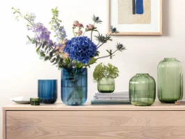 Green and blue glass vases in various shapes some with flowers in them standing on wood furniture with a picture on the wall behind them