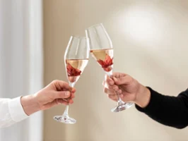 Two people toasting with La Divina glasses Against a neutral background, two people are raising a toast with Villeroy & Boch La Divina glasses, with a hibiscus flower in each one.