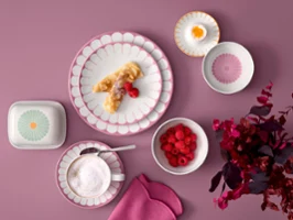 Fleur breakfast tableware set on pink background Overhead flat lay of Villeroy & Boch's Fleur pink porcelain plates with a crepe, a cup of cappuccino, a butter dish, and small bowls filled with fresh raspberries and a soft-boiled egg, set on a dusty pink background next to magenta foliage.