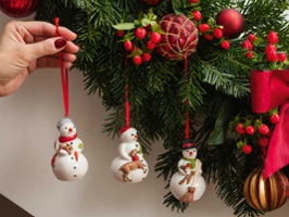 Mit der Hand hängen Schneemann-Ornamente an einen geschmückten Weihnachtsbaum mit roten Bändern, Beeren und Kugeln.
