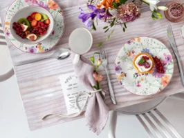 Ein mit Blumenmuster gedeckter Tisch mit Tellern, Besteck, einer Serviette, einem kleinen Dessert und einer Obstschale auf einer hellen Tischdecke.