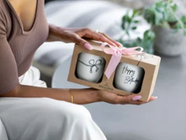 Une personne tient une boîte cadeau avec un ruban rose, à l’intérieur de laquelle se trouvent deux mugs « Happy Birthday ».