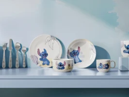 A set of plates and cups from the Disney Stitch collection featuring cartoon motifs are displayed on a light blue shelf with utensils.