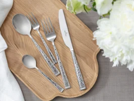 Vue de dessus des couverts Blacksmith – cuillère à soupe, fourchette de table, couteau de table, cuillère à café et fourchette à dessert – soigneusement disposés sur une planche en bois. Entourés de délicates décorations florales blanches. Une esthétique propre, naturelle et élégante.