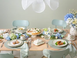 A pastel-themed dining table set for four, featuring plates of food, teacups, blue and white floral arrangements, and elegant tableware, creating a fresh and inviting atmosphere.