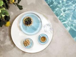 A round tray with tableware from the Perlemor Aqua collection A round tray with tableware from the Perlemor Aqua collection and a glass of water, at the edge of a pool.