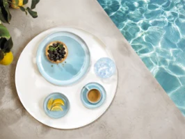A round tray with tableware from the Perlemor Aqua collection A round tray with tableware from the Perlemor Aqua collection and a glass of water, at the edge of a pool.