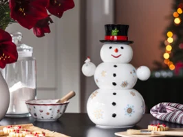 Ein Weihnachtsmann von Villeroy & Boch mit schwarzem Hut und roter Schleife steht auf einem Tisch neben einer Zuckerdose, einer Rührschüssel und roten Blumen. Im Hintergrund sind Weihnachtslichter verschwommen.