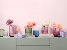 Una serie di vasi dai colori pastello della collezione Fleur, di diverse forme e dimensioni, con fiori rosa, bianchi e color pesca, disposti su uno scaffale con libri impilati e piccole scatole.