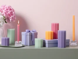 Una collezione di candele decorative colorate, della collezione Fleur, in diverse forme e tonalità pastello, è disposta su una superficie verde, con fiori rosa sullo sfondo davanti a una parete rosa chiaro.