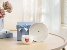 White mug from With Love with red heart motif, matching saucer, and blue gift box with bow on a wooden table by the window.