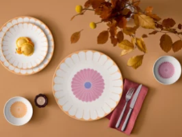 Flat lay di piatti e ciotole Fleur in calde tonalità gialle Vista dall'alto di piatti e ciotole in porcellana Fleur con vivaci motivi floreali a petalo su uno sfondo ocra caldo, decorati con posate su un tovagliolo rosso e rami autunnali.