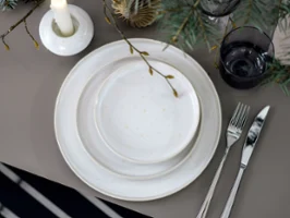 Eine festlich gedeckte Tafel, geschmückt mit Winter Glow Tellern, Besteck und einem Winter Glow Kerzenständer, umgeben von Tannenzweigen.