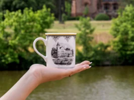 Audun cup in front of a river and trees A hand holding a decorative Audun cup with an illustration of a building in front of a river and trees.