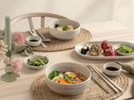A table set for two with Perlemor pieces A table set for two with bowls of ramen, sushi rolls, edamame, chopsticks and dipping sauces from the Perlemor collection on woven placemats.