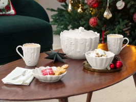 Ein geschmückter Weihnachtsbaum mit roten Ornamenten steht hinter einem Couchtisch, auf dem Tassen, Kekse und festliche Dekorationen in einem gemütlichen Wohnzimmer stehen.

