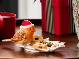 Toy's Fantasy bird Sur une assiette festive de Villeroy & Boch se trouvent une figurine d'oiseau avec un bonnet de Noël et quelques biscuits, à côté se trouvent un cadeau emballé et un vase en verre.