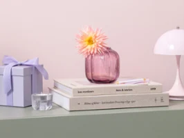 A small pink glass vase from Fleur with a flower stands on stacked books on a pastel green shelf.