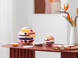 La petite boule miami and la boule miami arranged on a red table Colorful, multi-piece tableware sets from la petite boule miami, circular design objects in large and small sizes, stand as decoration next to a vase with abstract patterns and orange flowers on a red table.