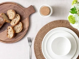 Ein gedeckter Tisch mit weißem Geschirr auf Rattan-Tischsets und einem Holztablett mit Brot
