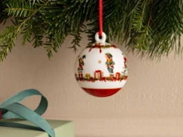 A white Christmas tree ornament featuring a festive scene by Villeroy & Boch hangs from a red string attached to a fir branch above a wrapped gift.
