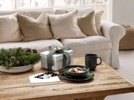 A wooden coffee table with a lave green a mug, snacks, plants, and paper in front of a white sofa with beige pillows.