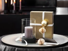 A Manufacture hurricane lamp with a candle next to a gift wrapped with a white box and ornaments.